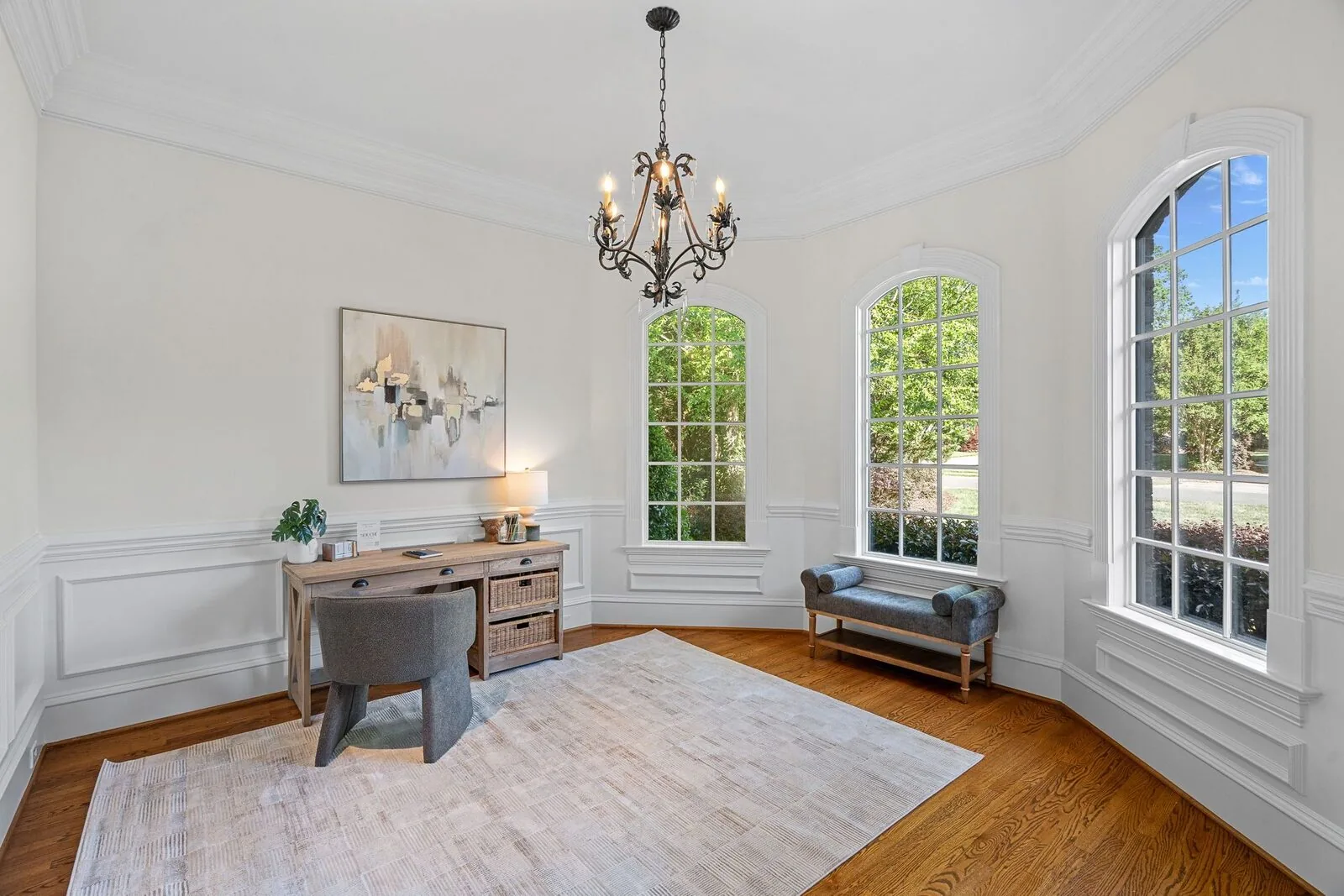Staged study with a writing desk, upholstered chair, artwork, and tall windows.